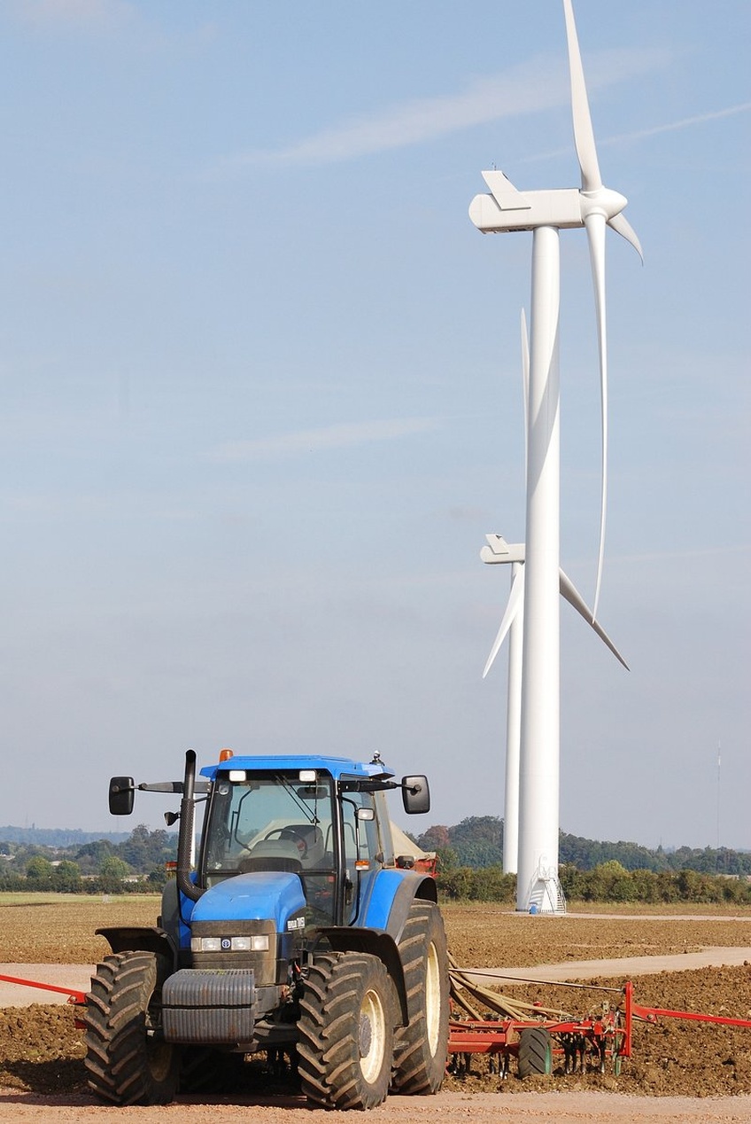 En modern blå traktor med röd utrustning arbetar på ett plöjt fält med vindkraftverk i bakgrunden.